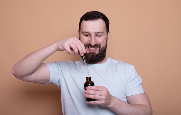 Bearded man holding pipette with beard oil on beige background