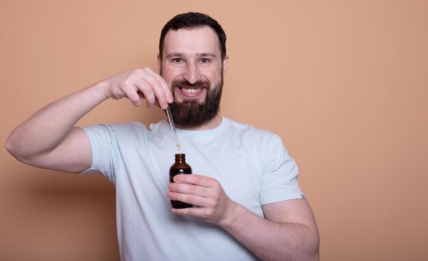 Bearded man holding pipette with beard oil on beige background