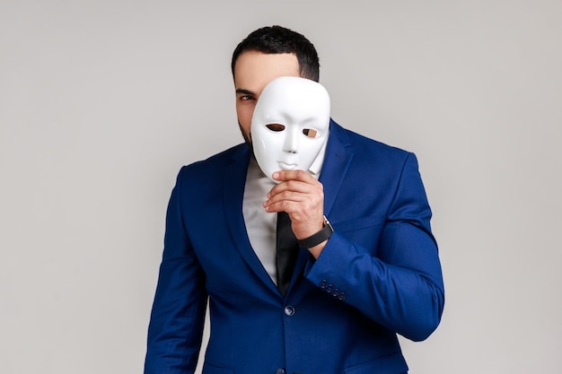 Bearded man holding white mask covering face standing with serious expression multiple personality disorder wearing official style suit Indoor studio shot isolated on gray background