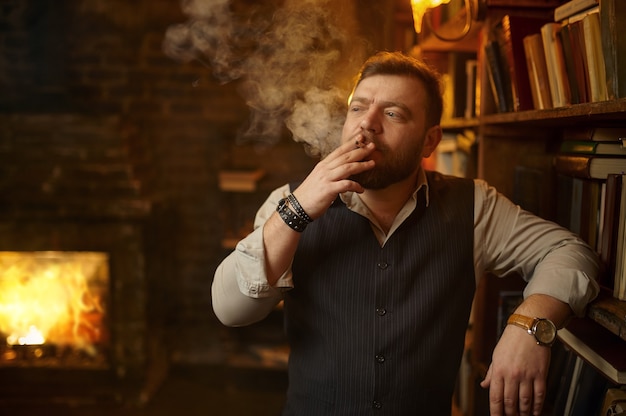 Bearded man holds ashtray and smokes cigarette, bookshelf and rich office interior on background. Tobacco smoking culture, specific flavor