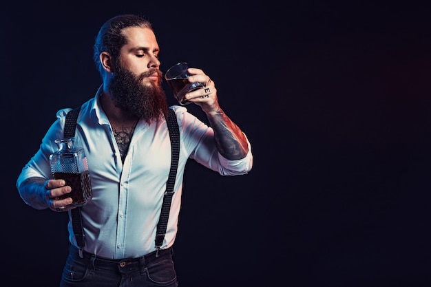 Bearded man holds bottle of whiskey in his hand and drinks dressed in white shirt studio shot