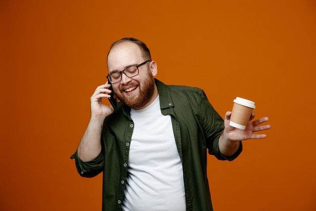写真 オレンジ色の背景の上に元気に立って笑っているコーヒーのカップを保持している携帯電話で話している眼鏡をかけてカジュアルな服を着たひげを生やした男