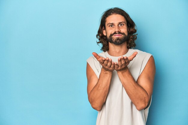 Foto bearded man in een tank top blauwe achtergrond vouwen lippen en het houden van handpalmen om lucht kus te sturen