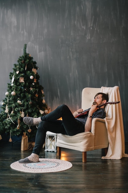 Photo bearded man is sitting comfortably in an armchair with a guitar in his hands near a christmas tree