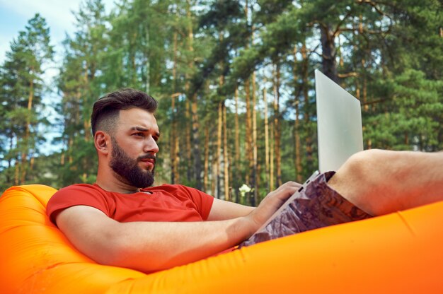 Bearded man is working and smiling with laptop at the park under the tree. happy freelancer is sitting and using app or website on the grass. work remotely in self-isolation mode