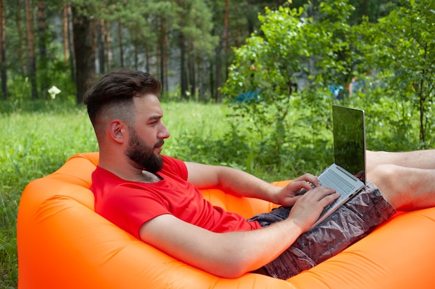 Bearded man is working and smiling with laptop at the park under the tree. Happy freelancer is