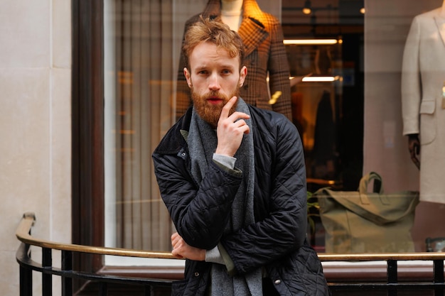 Bearded man in jacket and scarf posing outdoors near clothing store