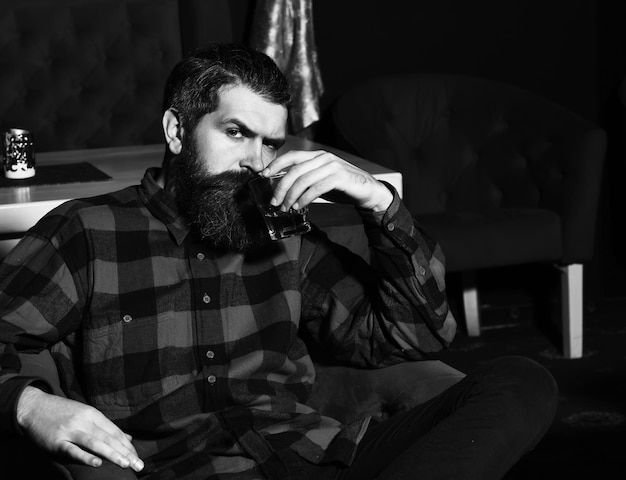 bearded man, long beard, brutal caucasian hipster with moustache holding glass with whiskey has serious face, unshaven guy with stylish hair in checkered shirt sitting in red chair, armchair