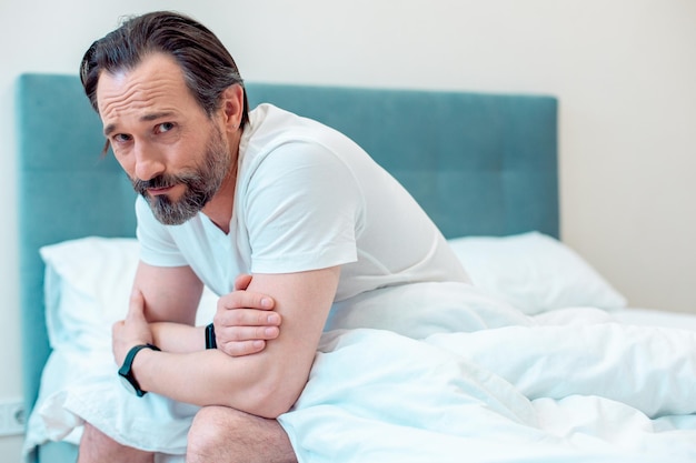 Bearded man looking shy while sitting in white tshirt on the\
bed alone with a blanket covering his legs