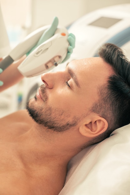 Bearded man lying with closed eyes and undergoing the photo rejuvenation procedure in modern clinic