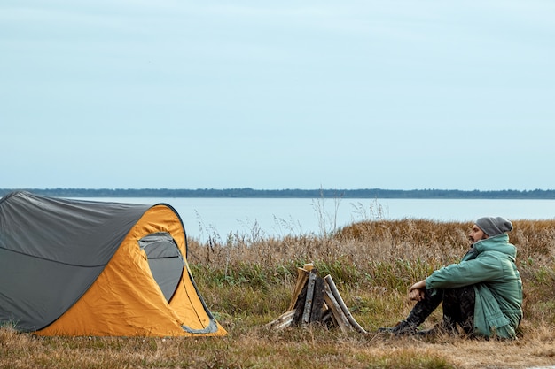 緑の自然と湖のキャンプテントの近くのひげを生やした男。旅行、観光、キャンプ。