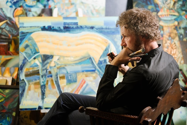 Бородатый художник в костюме сидит в галерее возле своих картин