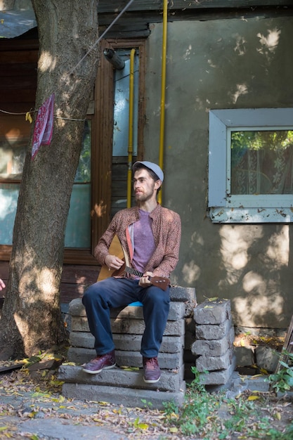 A bearded man playing balalaika sitting on the stairs of an old country house