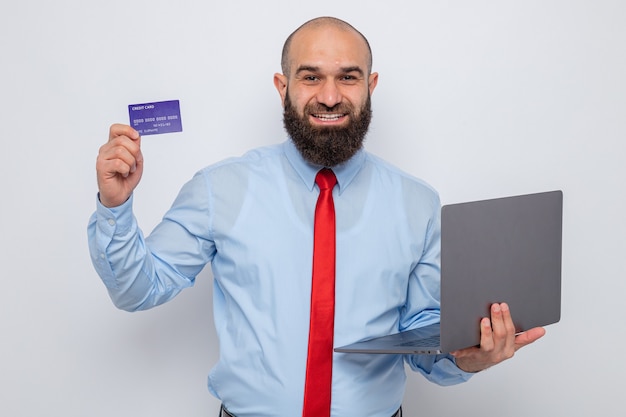 Bearded man in red tie and blue shirt holding laptop and credit card looking smiling cheerfully happy and positive