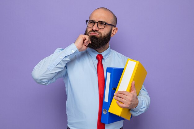 Bearded man in red tie and blue shirt wearing glasses holding office folders looking up puzzled standing over purple background