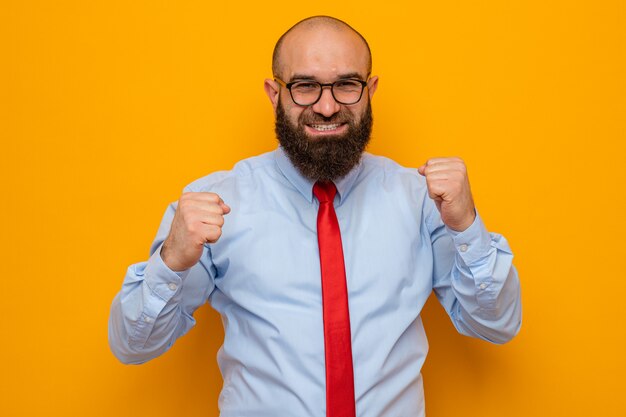 Bearded man in red tie and blue shirt wearing glasses looking happy and excited clenching fists