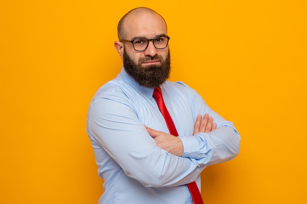 Bearded man in red tie and shirt wearing glasses looking at camera with serious confident expression with arms crossed standing over orange background