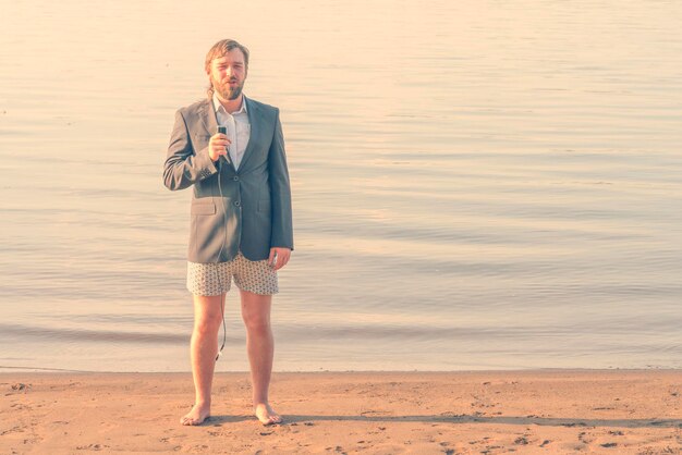 Bearded man reporter in a gray jacket and underwear with a microphone in his hand on the beach near the water Toned