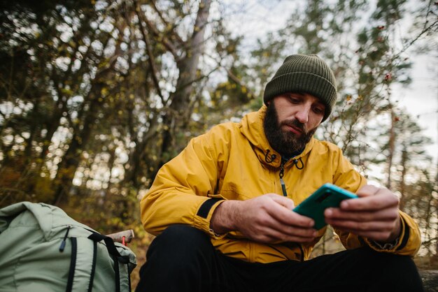 ひげを生やした男が森の真ん中で倒れた木の上で休んでいます 携帯電話を使って GPS を検索する男