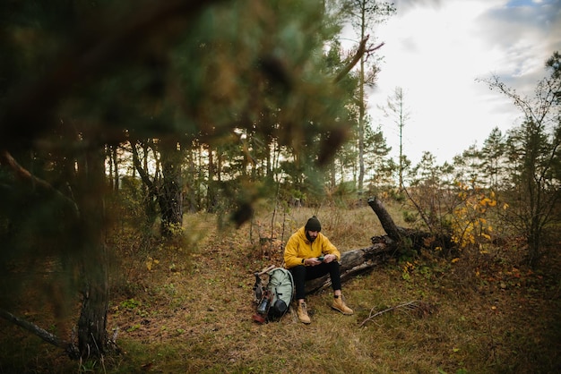 ひげを生やした男が森の真ん中で倒れた木に休む 携帯電話を使って GPS を検索する男