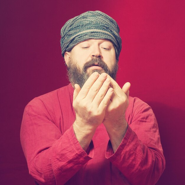 A bearded man in a shirt and a turban in a posture of prayer looks at his hands on a red background Toned