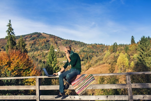 Bearded man sits on railing terrace and working on laptop, talks on phone, drinking tea. Freelancer hipster on vacation in mountains