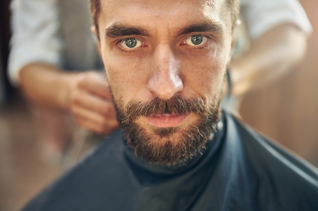 a bearded man sitting in an armchair and having a barber fix a cape around him