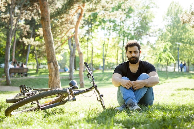 Bearded man sitting next to his bike on the ground in the park