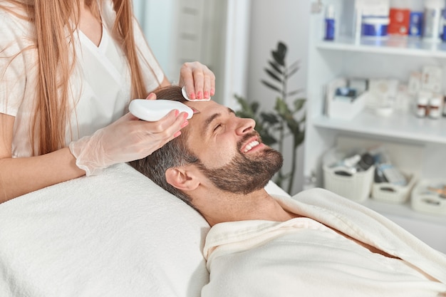 Bearded man smiling in professional beauty spa salon during ultrasonic facial cleansing procedure.