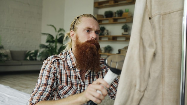 Bearded man steaming his jacket in bedroom at home