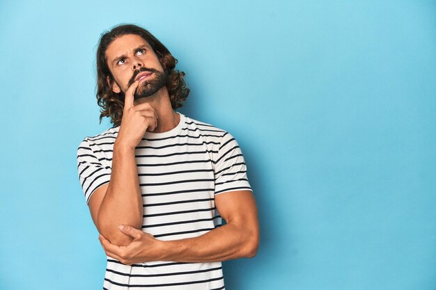 Foto uomo barbuto con una camicia a righe sullo sfondo blu rilassato pensando a qualcosa che guarda