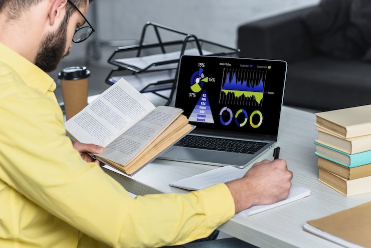 Photo bearded man studying with book near laptop with charts and graphs on screen in modern office