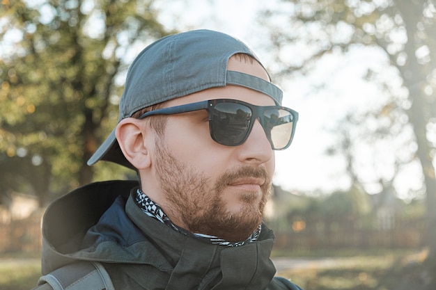 Bearded man in sunglasses and a baseball cap on the background of trees and nature on a sunny autumn day.