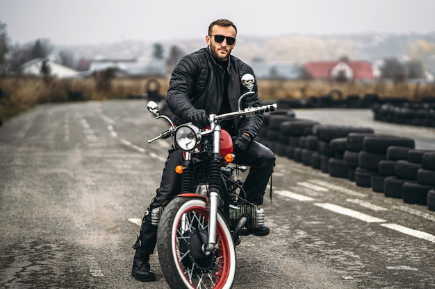 Uomo barbuto in occhiali da sole e giacca di pelle seduto su una moto sulla strada.