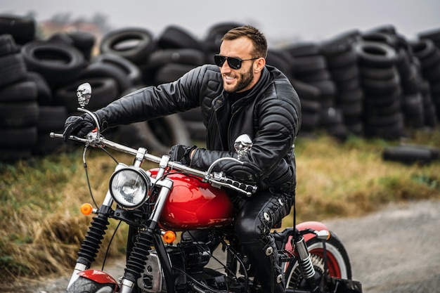 Bearded man in sunglasses and leather jacket smiling while sitting on a red motorcycle on the road.