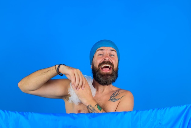 Bearded man take a shower. Bath time. Body washing. Hair care. Man with bath hat on head. Bath sponge.