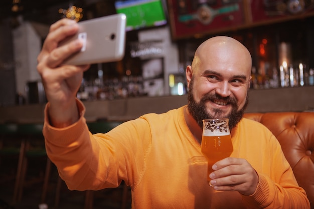 Бородатый мужчина берет селфи со стаканом пива в руке, используя смартфон в пабе