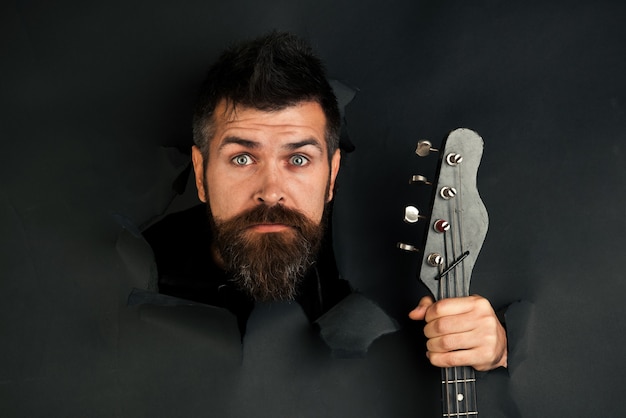 Bearded man through hole in black paper holds guitar in hand surprised man with beard and mustache