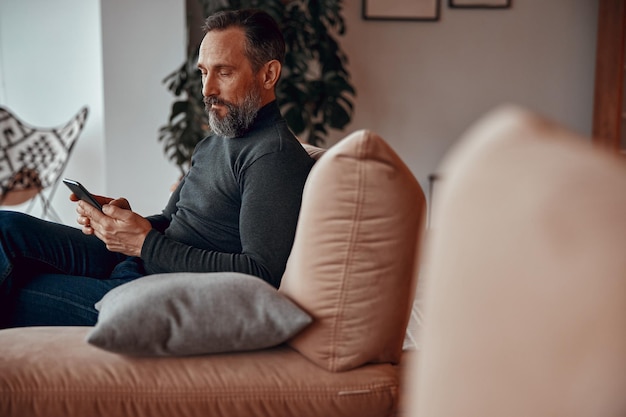 Photo bearded man typing message on phone in cosy armchair