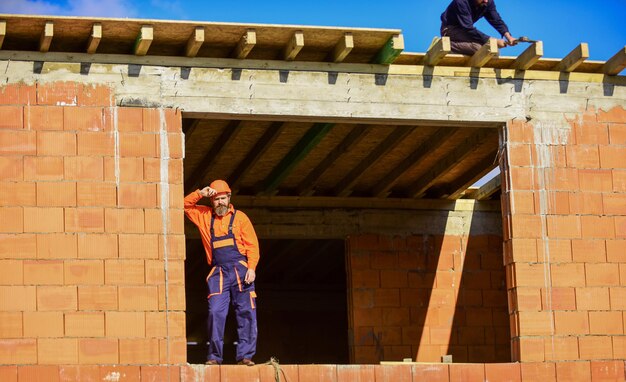 Bearded man in uniform and hard hat repairman engineer wear helmet on site tired builder relax construction worker smoking cigarette building is under construction