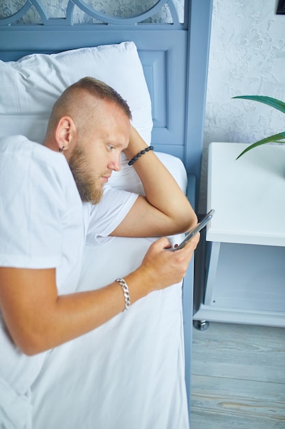 Uomo barbuto che utilizza lo smartphone al mattino nell'accogliente sdraiato sul letto