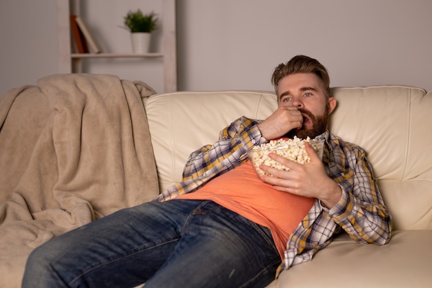 Bearded man watching film or sport games TV eating popcorn in house at night. Cinema, championship and leisure concept.