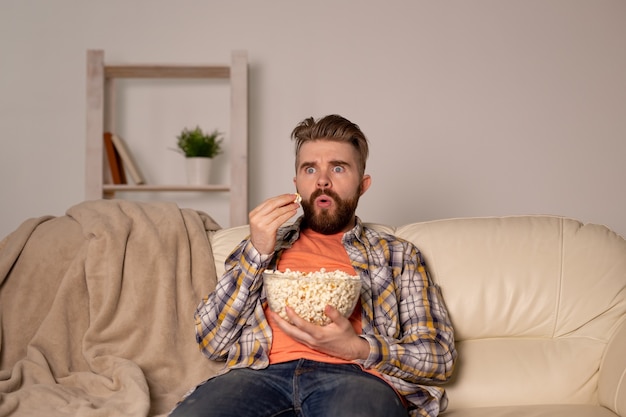 Бородатый мужчина смотрит фильм или спортивные игры по телевизору, ест попкорн в доме ночью. Кино, чемпионат