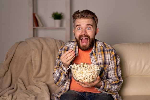Bearded man watching film or sport games TV eating popcorn in house at night. Cinema, championship