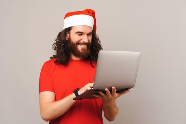 Bearded man wearing Christmas hat is holding a laptop while typing on it