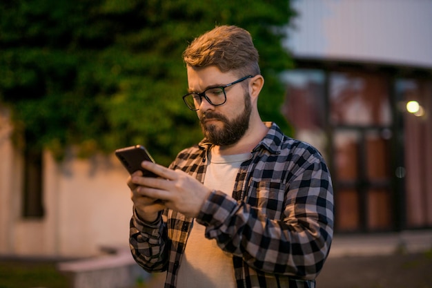 Бородатый мужчина в очках держит прокрутку текстовых сообщений в своем мобильном телефоне ночью, уличный парень, кэл.
