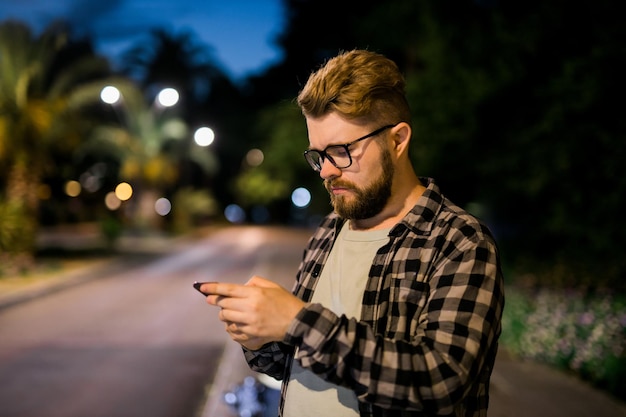 眼鏡をかけているひげを生やした男は、夜の街の男calで彼の携帯電話でスクロールテキストメッセージを保持しています