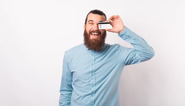 Bearded man wearing a light blue shirt is covering one eye with his credit card.