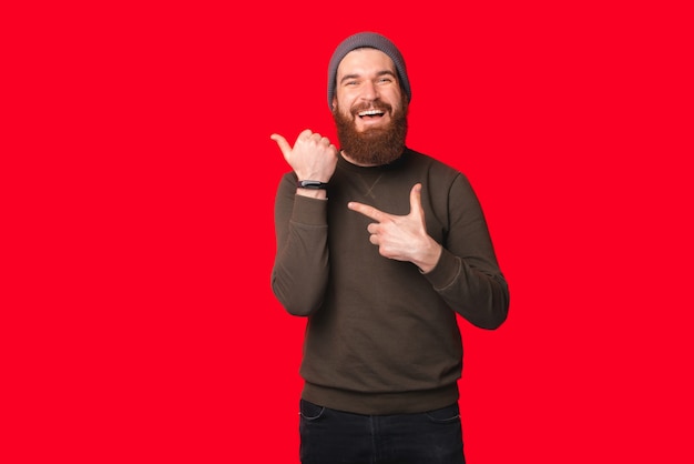 Bearded man wearing warm hat and sweater is pointing at his wrist watch.