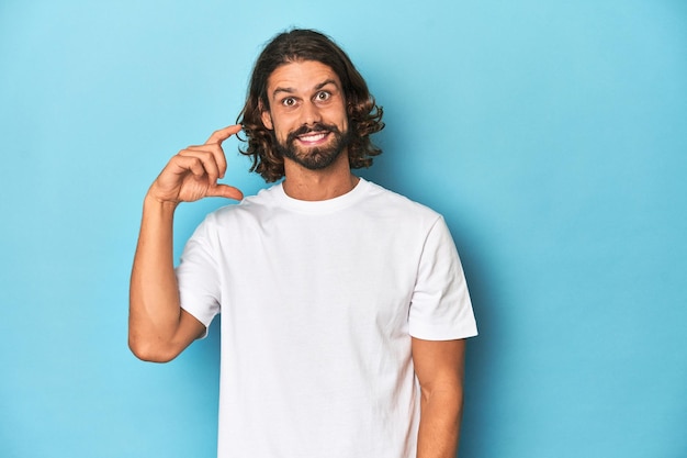 Foto uomo barbuto con una camicia bianca che tiene qualcosa di piccolo con le dita dell'indice sorridente e sicuro